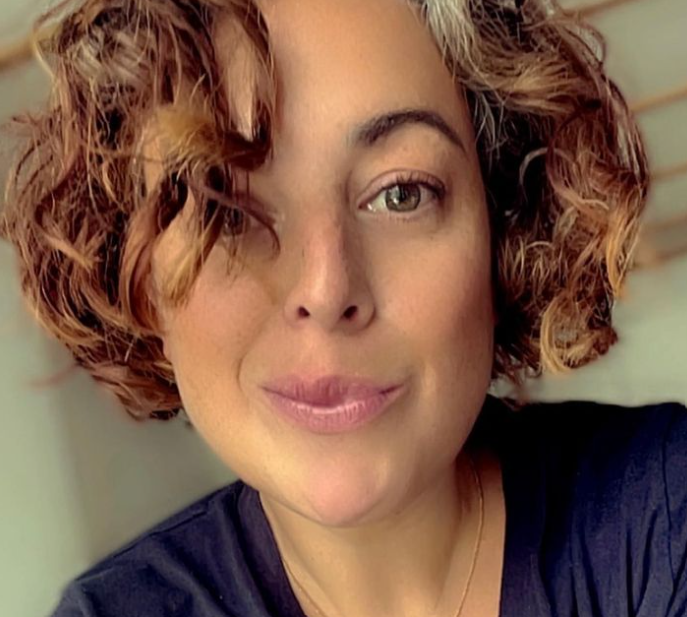 Porträt von Jule, lächelnd mit lockigem French-Bob, marineblauem T-Shirt und goldener zarter Kette. Nur die Schultern und der Hals sind sichtbar, der Hintergrund ist weichgezeichnet.