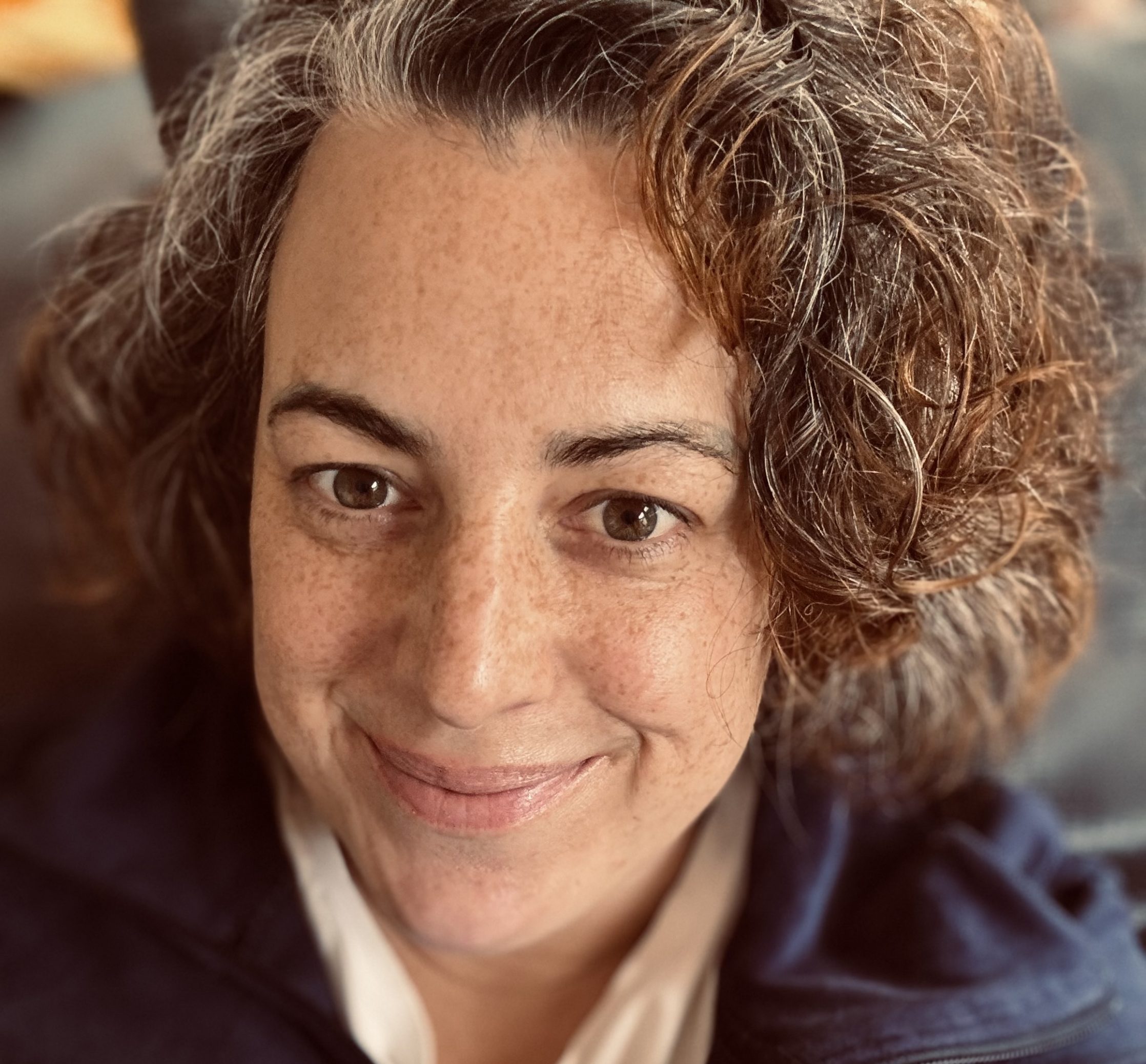 Nahaufnahme von Jule mit lockigen Haaren und Sommersprossen, lächelnd in einer blauen Jacke mit Stehkragen und beigefarbenem T-Shirt. Der Hintergrund ist weichgezeichnet. Das Bild steht für Achtsamkeit und den Weg zu ganzheitlicher Gesundheit.