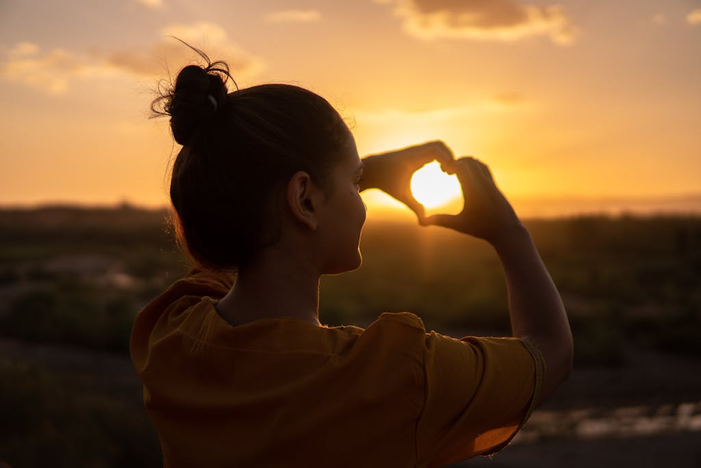 Eine junge Frau, Rückansciht, die glücklich und zufrieden mit ihren Händen ein HErzsymbol formt; Sonnenuntergang