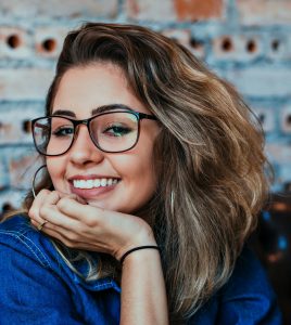 Porträt einer jungen Frau mit Brille und in Denim, die in einem Innenraum lächelt. Sie strahlt eine freundliche und einladende Atmosphäre aus, mit einem natürlichen Lächeln, das ihre Persönlichkeit unterstreicht. Die Frau trägt ein klassisches Denim-Outfit, das ihre entspannte Haltung im Raum widerspiegelt. Der Hintergrund ist weich und neutral gehalten, wodurch der Fokus auf ihr Lächeln und ihre positive Ausstrahlung gelegt wird.