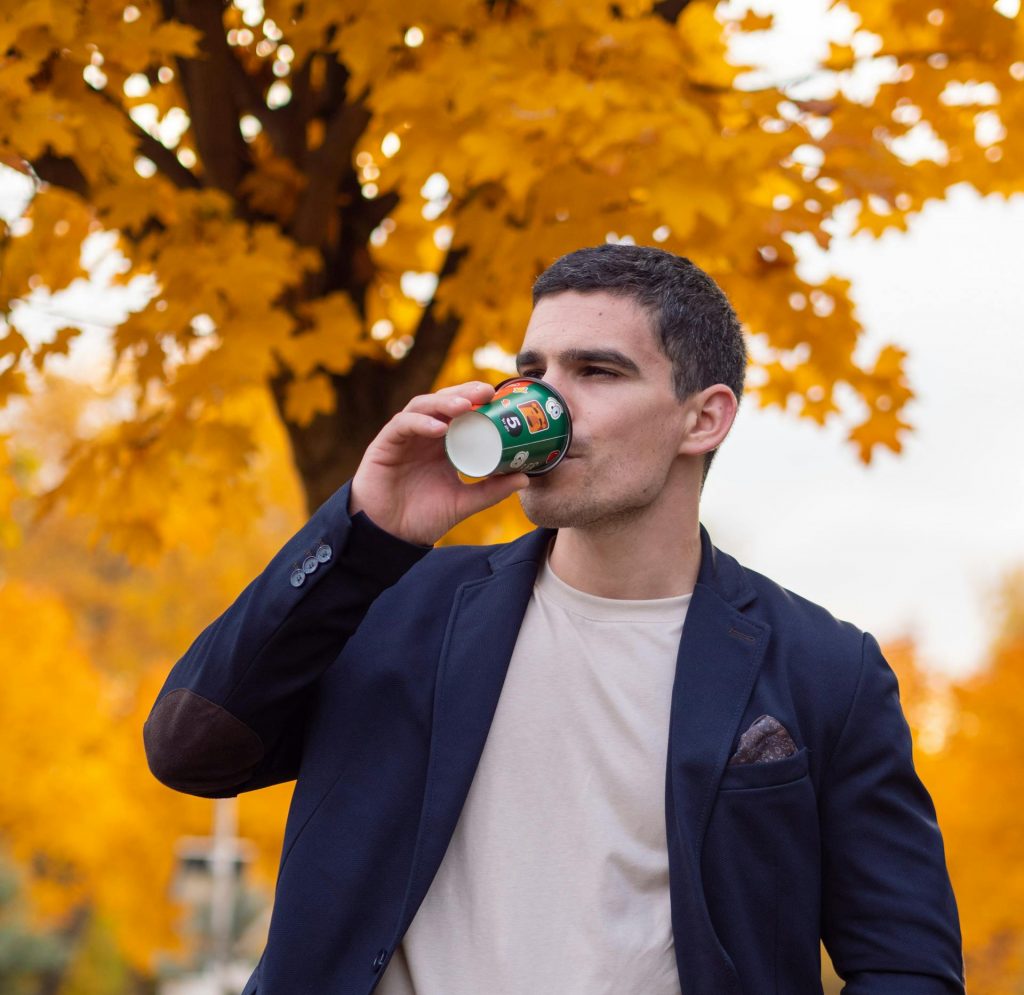Aufnahme eines Mannes bis etwa zum Bauchnabel, in dunkelblauem Anzug und weißem T-Shirt. Er steht vor einem Baum mit gelbem Herbstlaub und trinkt aus einem Becher. Der Hintergrund ist weichgezeichnet.