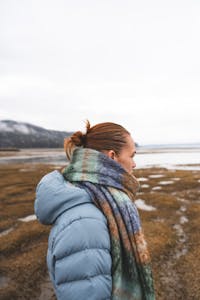 Seitliche Ansicht einer Frau in blauer Jacke und Schal, die an einem nebligen Strand steht. Die Szene vermittelt einen ruhigen Moment inmitten der Natur, mit sanften Nebelschwaden und einer friedlichen Atmosphäre.