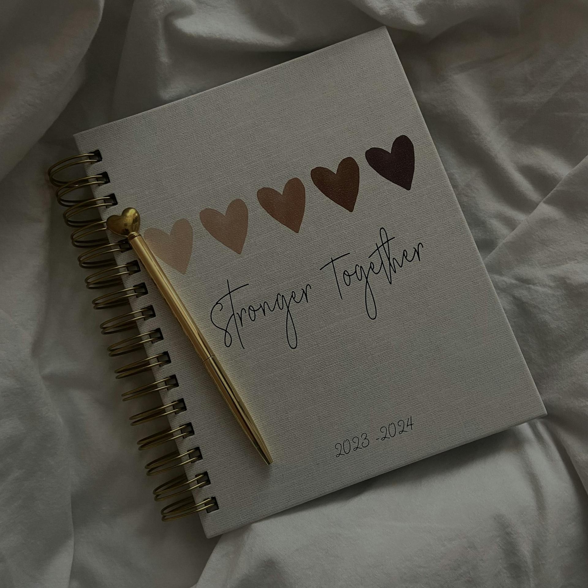 Spiralnotizbuch und Stift liegen auf einem Bett, darauf 5 Herzen von beige bis dunkelbraun, dazu der Text Stronger Together in einer Schmuckschrift