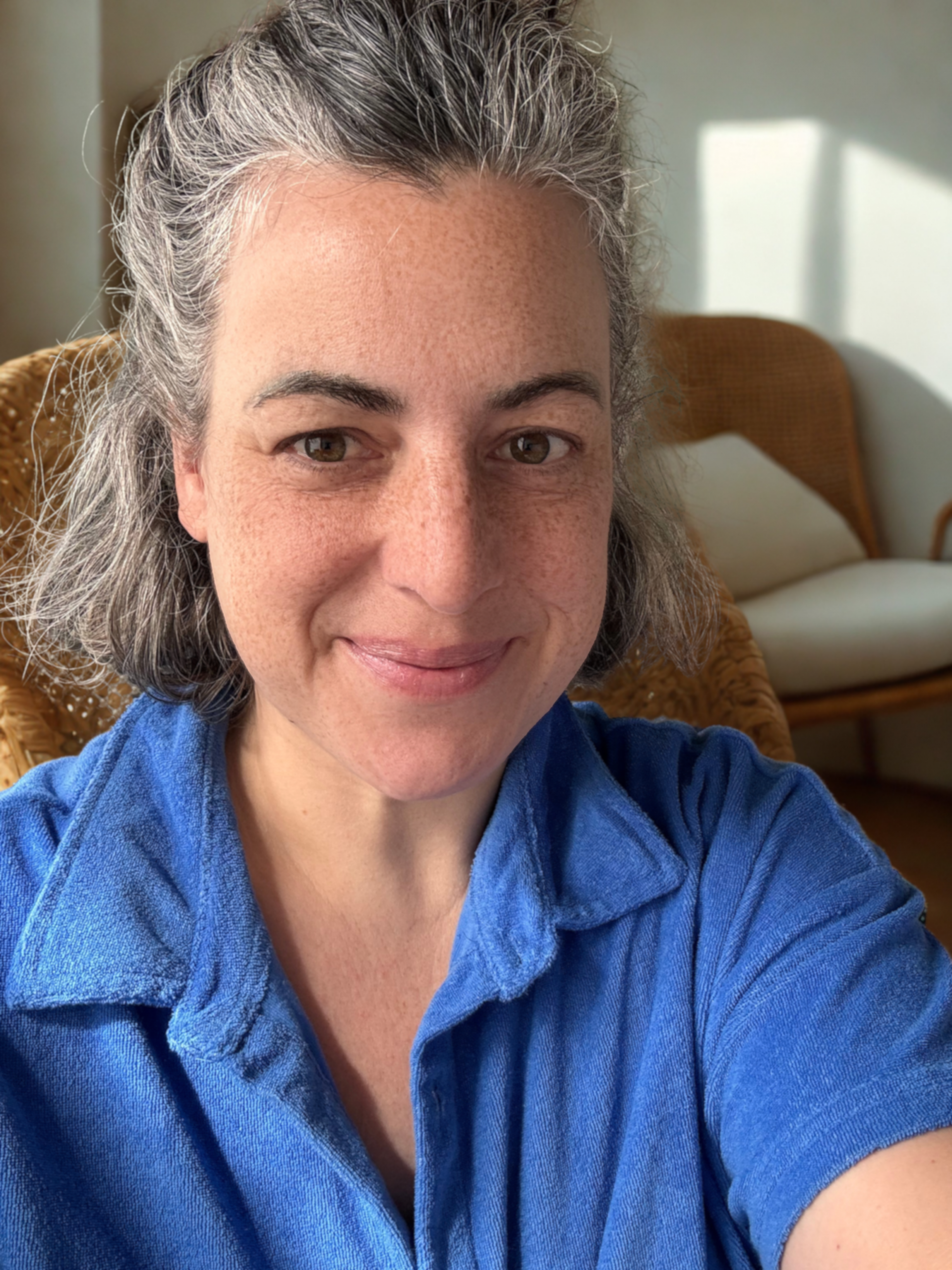 Jule, freundlich lächelnd im königsblauen Polo-Shirt, mit Half-Bun; im Hintergrund ein Rattanstuhl mit beigefarbenen Kissen.