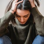 Junge Frau mit dunklem Haar in Alltagskleidung sitzt vor einer weißen Wand und hält den Kopf in den Händen. Ihre Haltung drückt Überforderung und emotionale Anspannung aus – ein starkes Bild für innere Krisen und psychische Belastung. Es symbolisiert, wie wichtig achtsame Begleitung und Selbstfürsorge in schwierigen Lebensphasen sind.