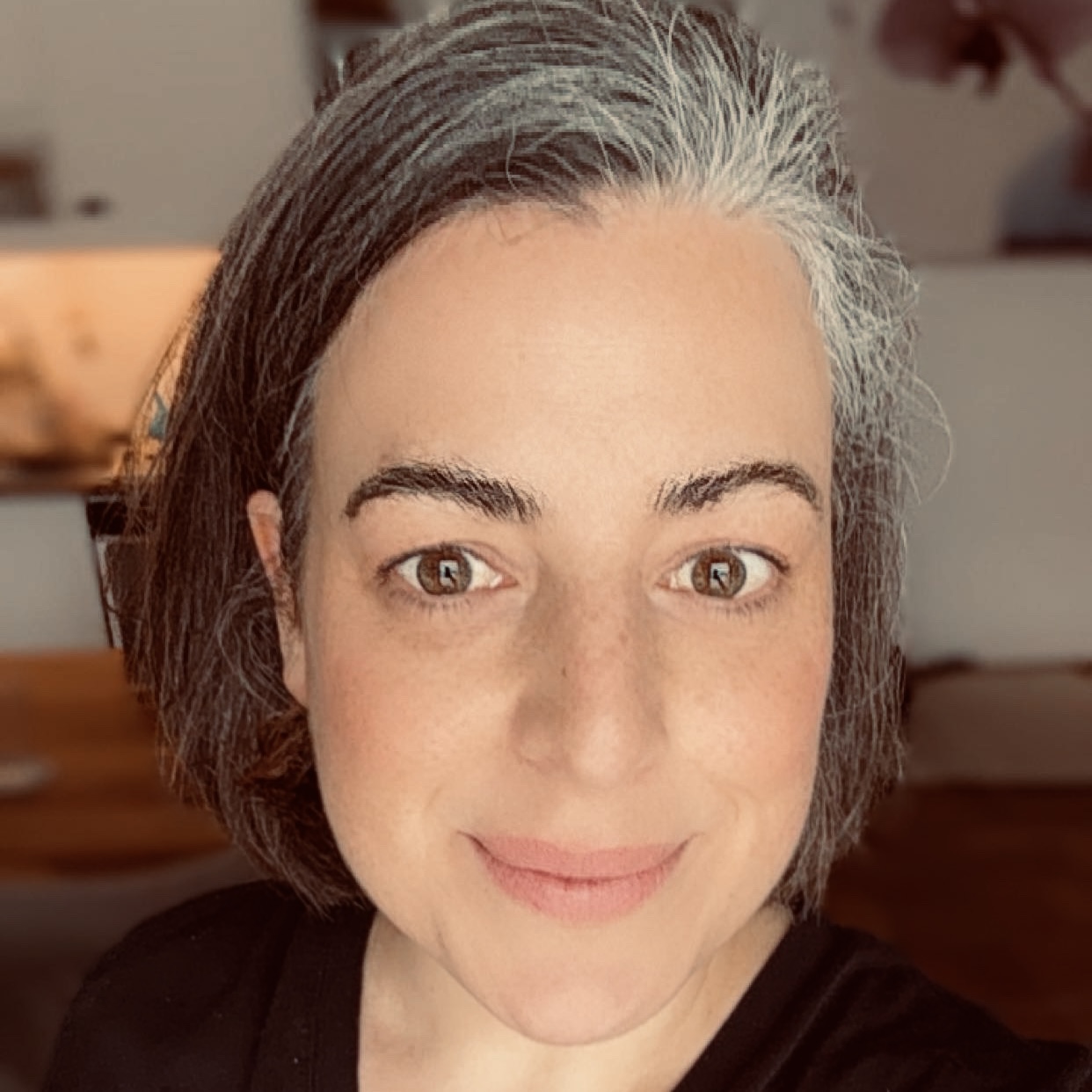 Porträt von Jule, lächelnd mit French-Bob, blauem T-Shirt und im Hintergrund ist ein Arbeitsraum verschwommen erkennbar.